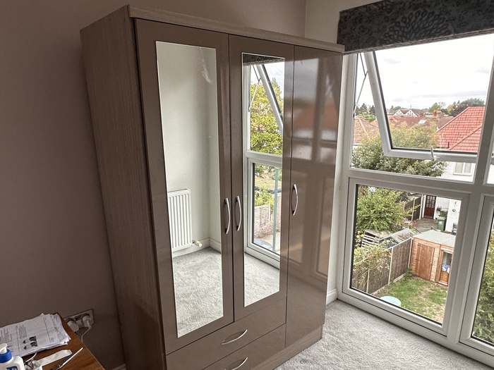 A three-door mirrored wardrobe assembled and set against the wall of a bay-window bedroom.